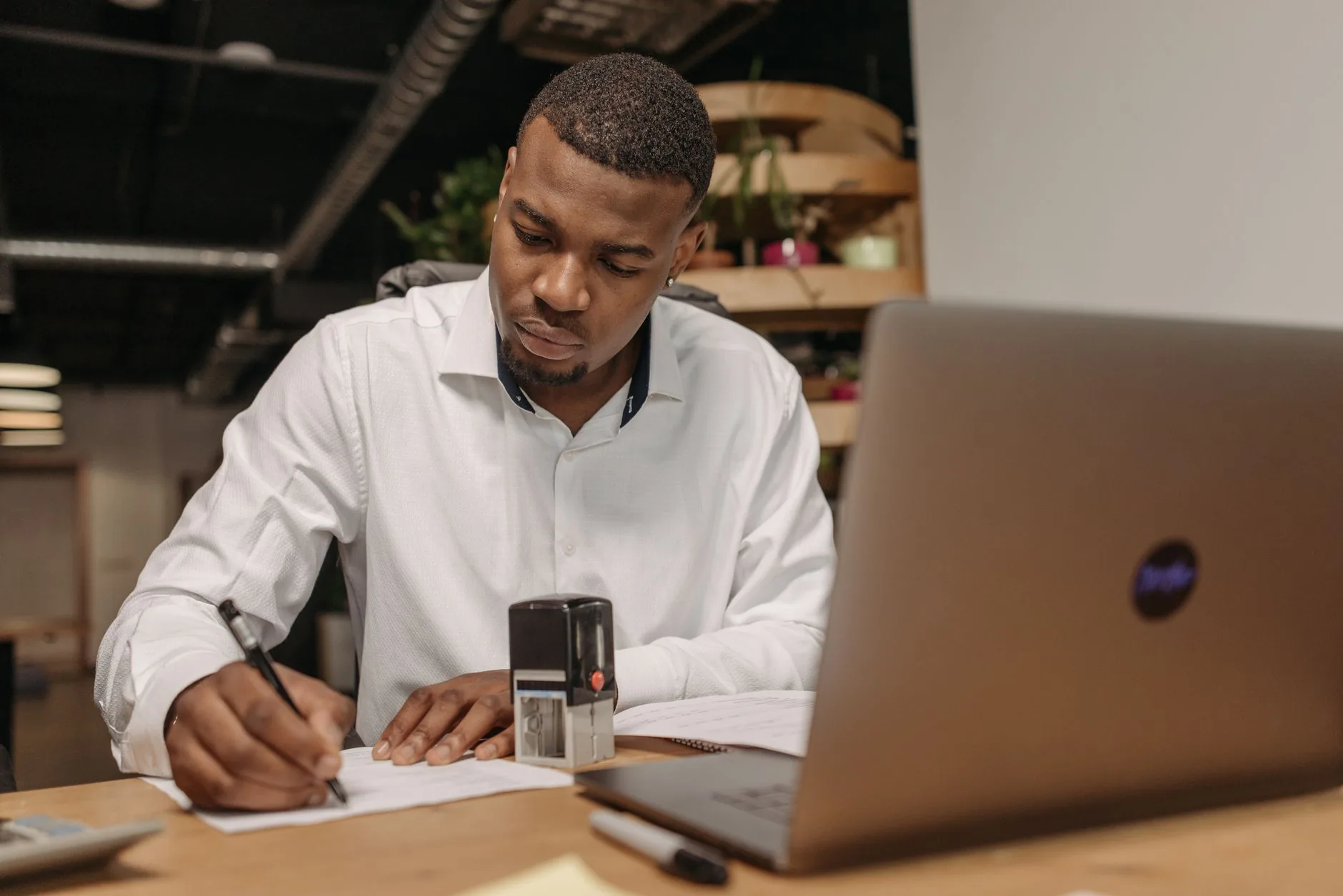 Un homme en chemise blanche écrit sur des documents à son bureau avec un ordinateur portable